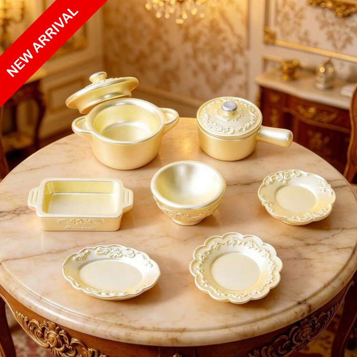 7-piece miniature gold cookware and tableware set displayed on a round marble table, featuring pots, a baking dish, a bowl, and decorative plates