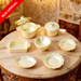 7-piece miniature gold cookware and tableware set displayed on a round marble table, featuring pots, a baking dish, a bowl, and decorative plates