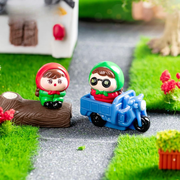 Miniature figurines set of 3 featuring a character in a red hood sitting on a log and another in a green hood driving a blue toy scooter amidst artificial grass and garden decor.