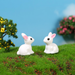 Two small glossy white miniature rabbits with pink ears facing each other on a green moss landscape with miniature trees in the background