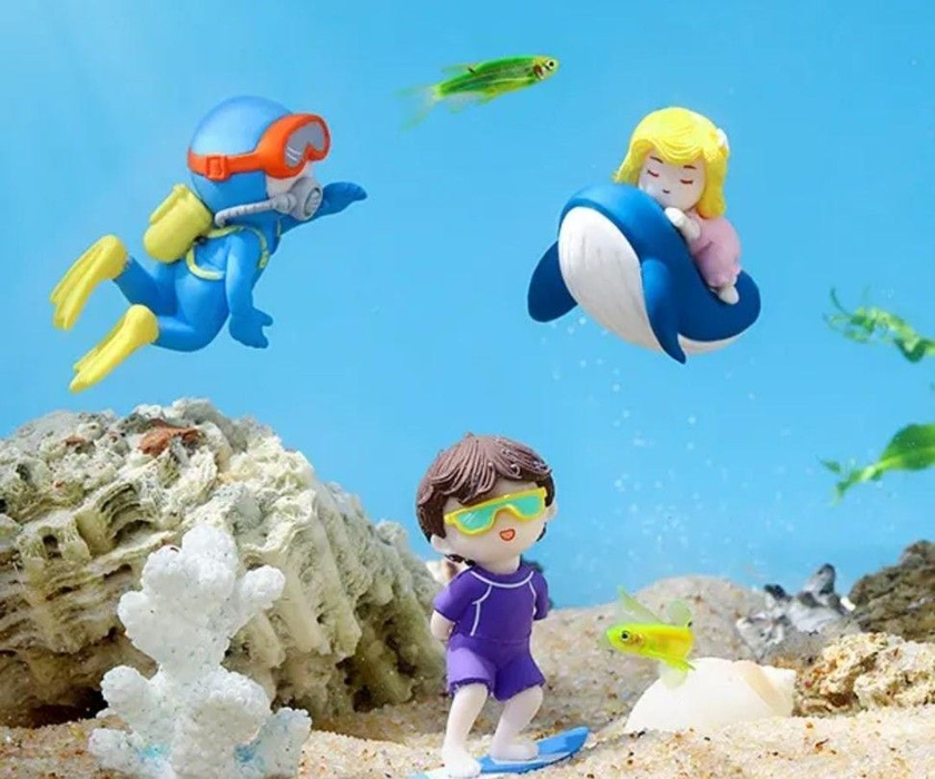 Set of 3 ocean miniature figurines featuring a scuba diver, a girl on a whale, and a surfer boy placed on sand.