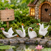 Set of 4 miniature white resin swan figurines with open wings placed by a fairy garden pond near a tiny cottage.
