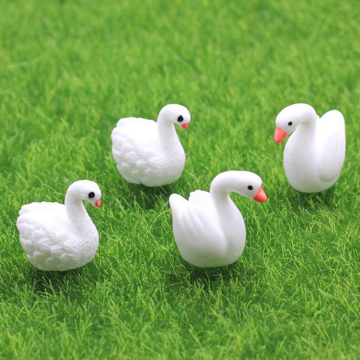 Four tiny white resin swan figurines with orange beaks arranged on a vibrant green artificial grass background.