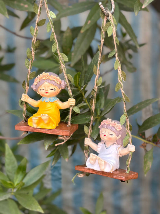 Two miniature fairy girl figurines with flower crowns sitting on wooden swings suspended by leaf-entwined twine in a garden setting.