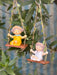 Two miniature fairy girl figurines with flower crowns sitting on wooden swings suspended by leaf-entwined twine in a garden setting.